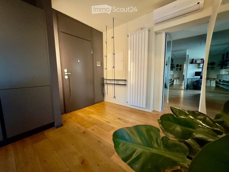 A modern hallway with wooden flooring, white walls, and a grey wardrobe on the left. A radiator is mounted on the wall, and a large green leafy plant is visible in the foreground. An open doorway leads to another room with visible furniture.