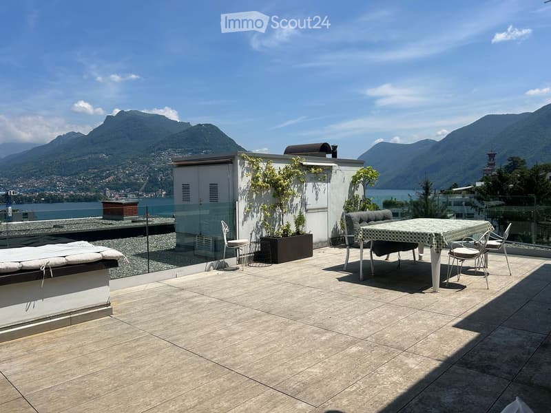 Roof terrace with tiled floor, outdoor furniture including a table, chairs, and a sofa, overlooking a scenic lake and mountain view.