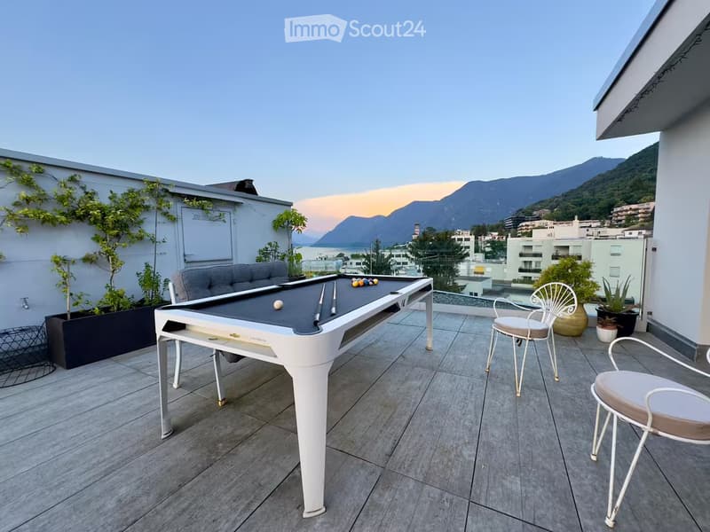 A rooftop terrace with a modern design, featuring a sofa, a white table with a pool setup, two white chairs, and several potted plants. The terrace overlooks a scenic view of mountains and a body of water.
