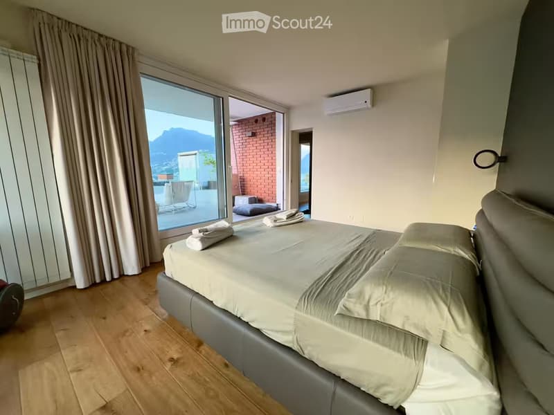 Modern bedroom with a large bed, pillows, and a blanket. The room features wooden flooring, large windows with a scenic mountain view, and neatly folded towels on the windowsill.