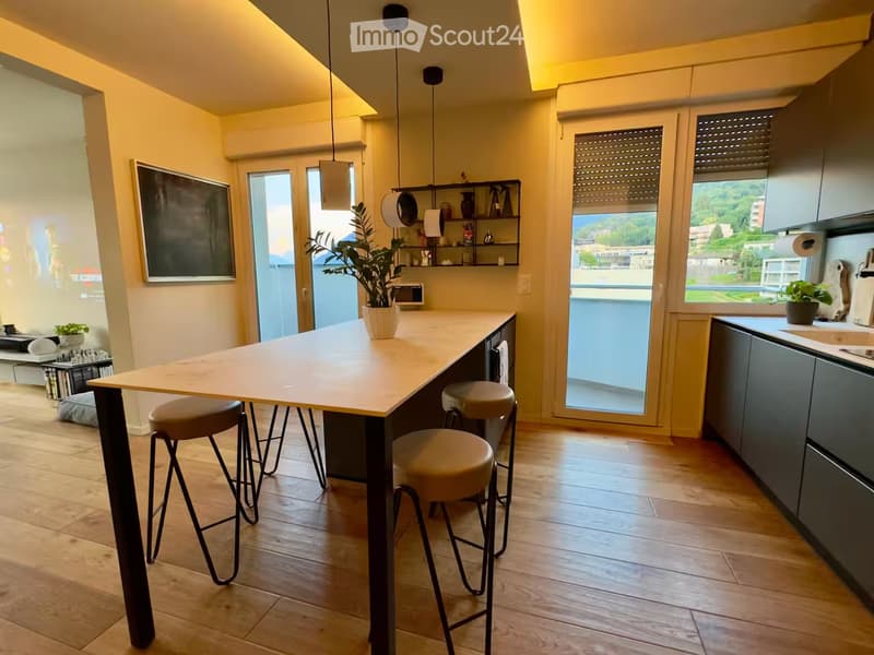 Modern kitchen with a central island, four stools, dark grey cabinets, a sink, and a view of greenery through large windows.