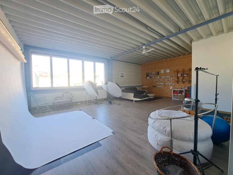 A spacious, well-lit room with wooden flooring, large windows, a sofa, a chair, a round table, a ball, a basket, and shelves with various items.