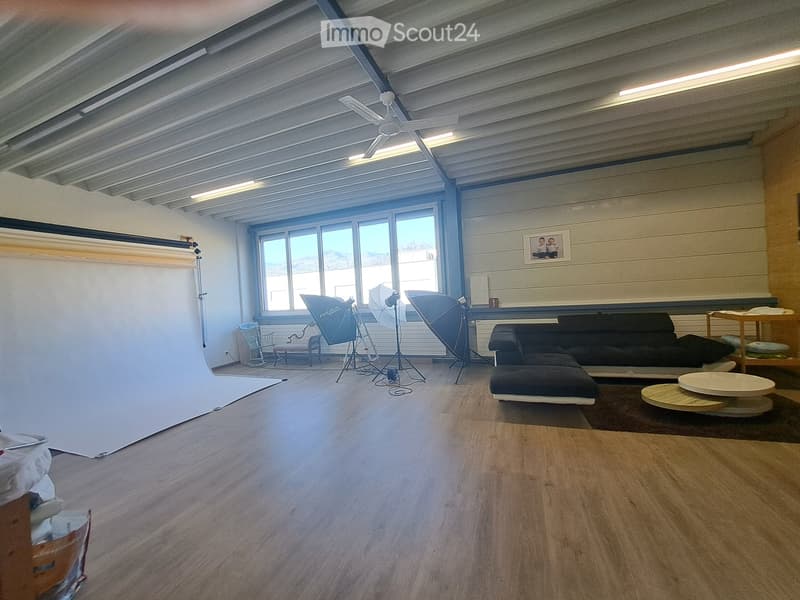 A spacious, well-lit room with wooden flooring, large windows, a sofa, coffee table, and photography equipment including umbrellas and tripods.