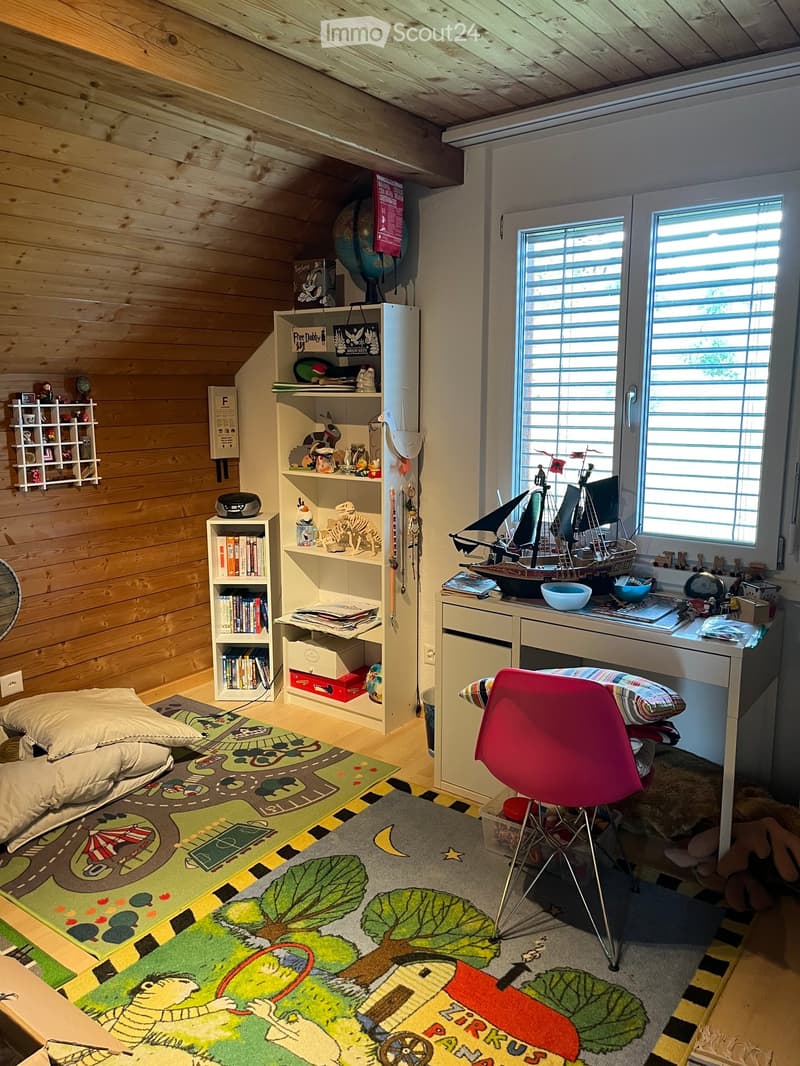 Cozy children's bedroom with wooden walls and ceiling, colorful rugs, a red chair, a desk with a toy ship, and shelves filled with books and toys.