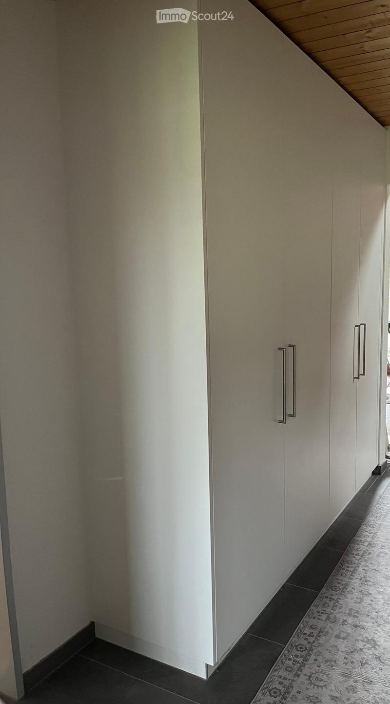 A narrow hallway with white walls, dark tiled flooring, and a patterned rug. The hallway features built-in white cabinets with sleek metallic handles on the right side.