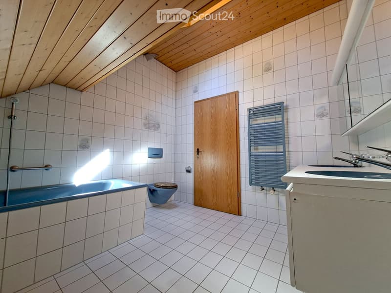 Bathroom with a blue bathtub, toilet, sink, mirror, and radiator. The room features white tiled walls and floor, a wooden door, and a slanted wooden ceiling.
