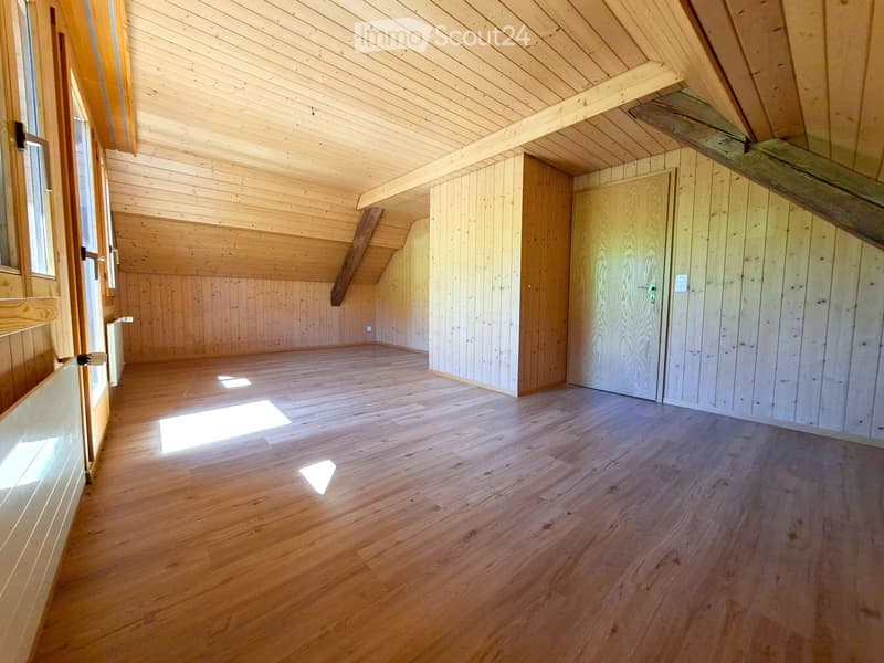 An empty attic space with wooden walls, ceiling beams, and hardwood flooring. Natural light streams in from a window on the left side.
