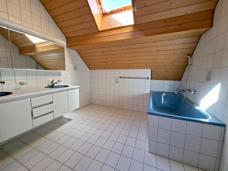 Bright bathroom with white tiled walls and floor, a blue bathtub, a double sink vanity with mirrors, and a skylight window providing natural light.