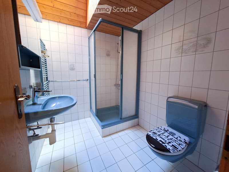 Bathroom with white tiled walls and floor, featuring a sink, toilet, and a glass-enclosed shower with a wooden ceiling.