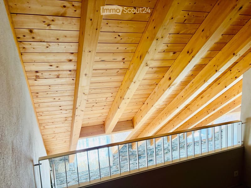 A modern hallway with exposed wooden beams and a sleek metal railing leading to a view of the outdoors.
