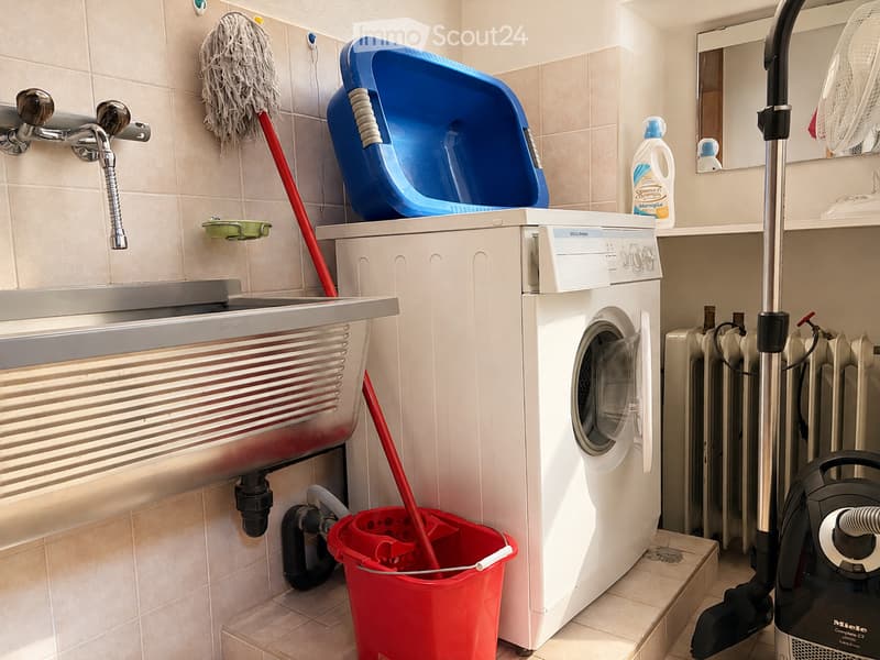 Bathroom with a washing machine, sink, mop, mop bucket, radiator, vacuum cleaner, fan, and a bottle on the counter.