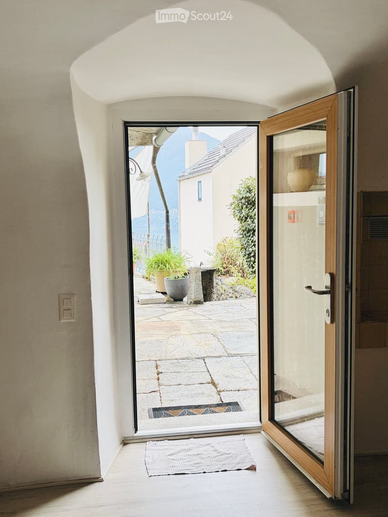 A narrow hallway with a sliding glass door leading to a stone-paved outdoor area, featuring a small white doormat inside and a view of a garden and distant mountains.