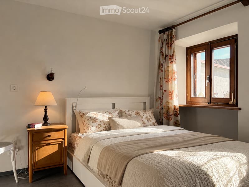 Cozy bedroom with a white bed, patterned pillows, a nightstand with a lamp, and a window with floral curtains.