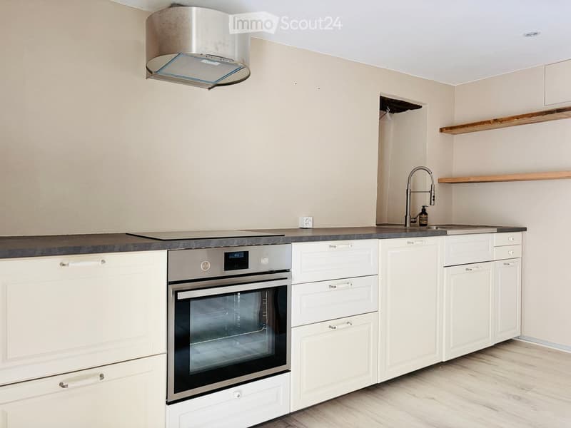 Modern kitchen with white cabinets, stainless steel oven, sink, and range hood. Wooden shelves and a sleek faucet are visible.