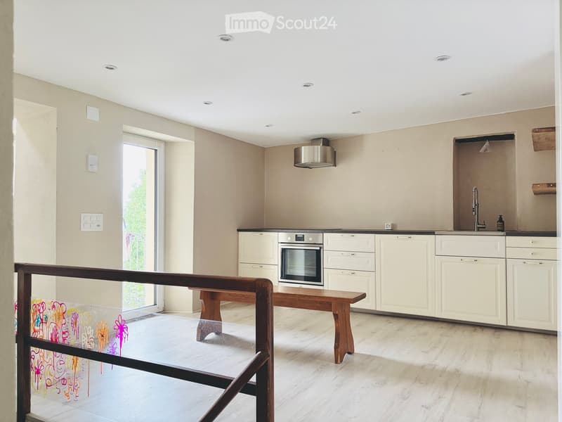 Modern kitchen with white cabinets, stainless steel oven, wooden bench, and a large window with a view of greenery.