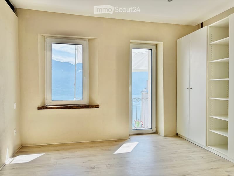 Empty room with light-colored walls, wooden flooring, a window with a scenic view, a door leading to a balcony, and built-in white shelving units.