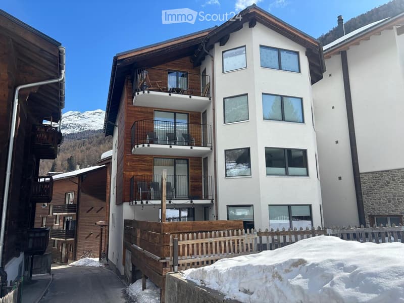 Multi-story residential building with white walls, wooden accents, multiple balconies, and snow-covered ground.