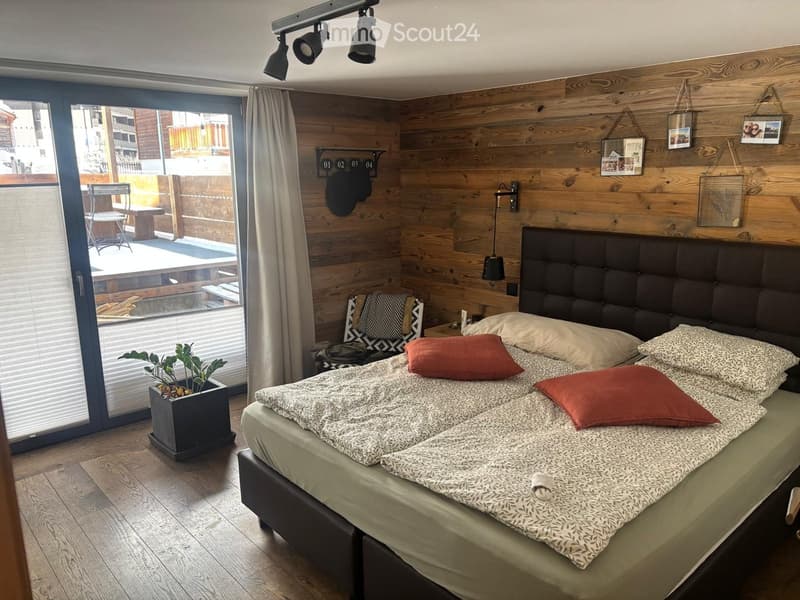 Modern bedroom with wooden walls, large bed with patterned bedding, nightstand, chair, and a small plant near the window. The room features a balcony with outdoor furniture visible through the large window.