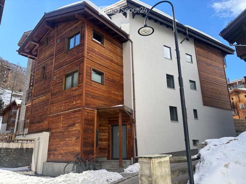 A two-story wooden house with white walls, multiple windows, and a snow-covered ground. A bicycle is parked near the entrance.