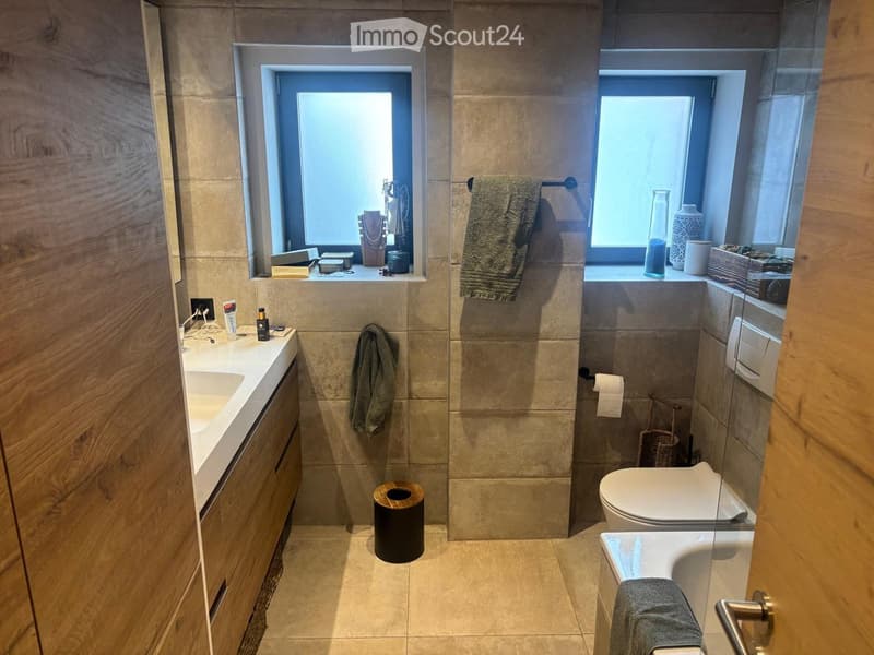 Modern bathroom with beige tiles, double sink, toilet, bathtub, and decorative items on windowsills.