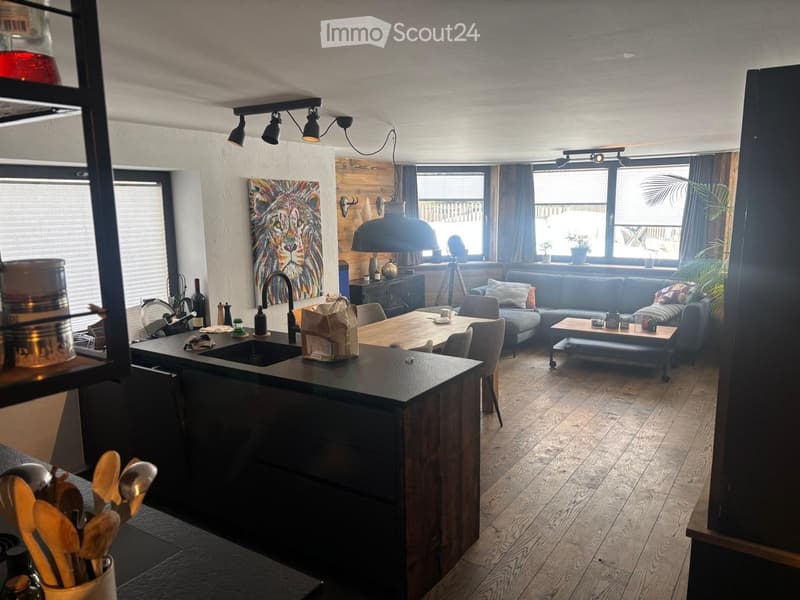 Open-plan living and kitchen area with wooden flooring, grey sofa, coffee table, kitchen island, sink, and large windows with curtains.