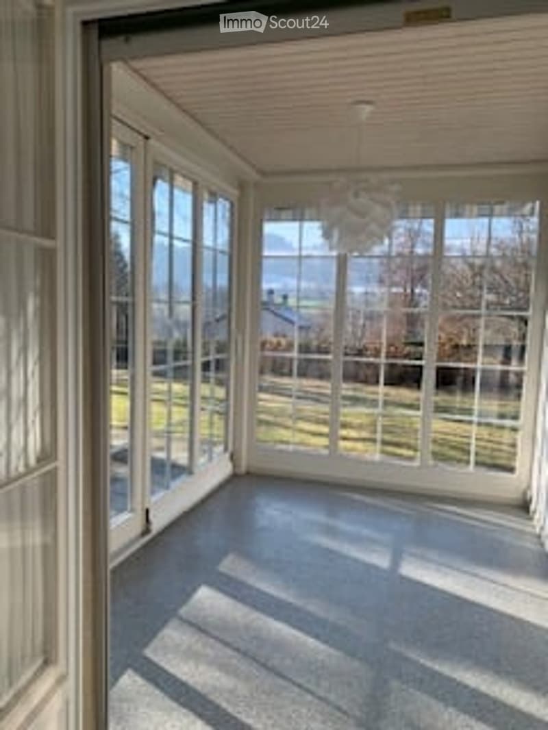 Bright, empty room with large windows and a view of a green outdoor area. The room has a smooth, light-colored floor and a simple ceiling light fixture.