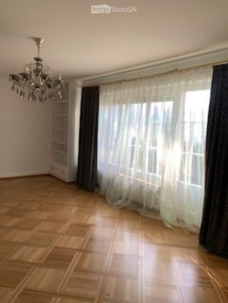 Empty room with wooden flooring, large window with white curtains, dark grey curtain, and a chandelier hanging from the ceiling.