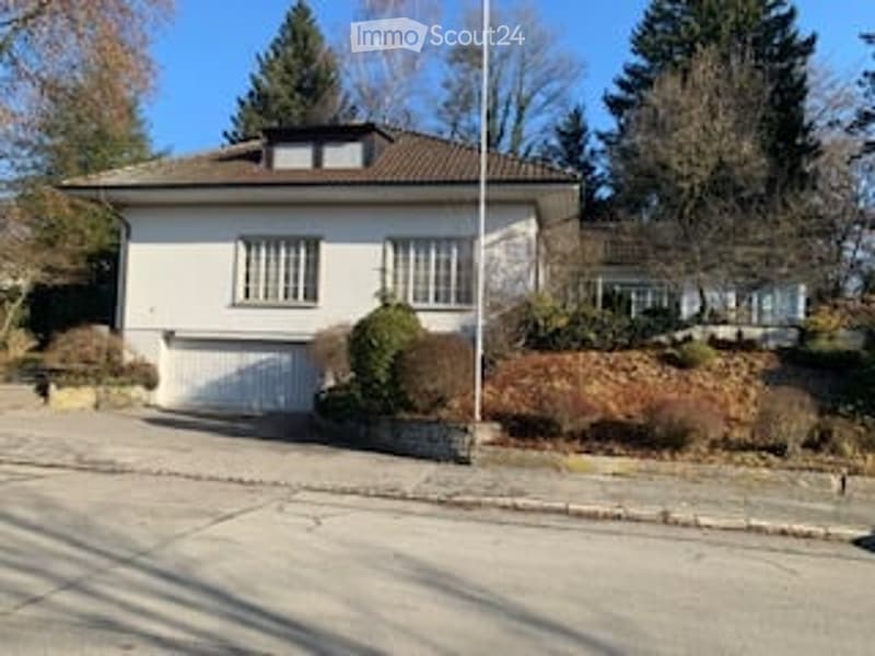 Single-story house with a white exterior, brown roof, and a garage. The house features multiple windows and is surrounded by a small garden and trees.