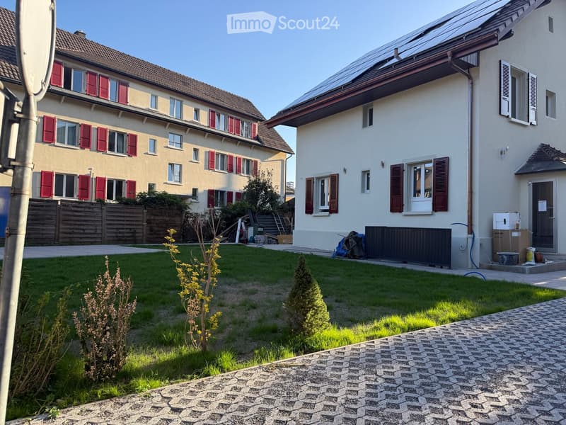 Well-maintained garden with green lawn, small plants, and a paved pathway leading to the house entrance. The house features light-colored walls, red shutters, and a modern design with solar panels on the roof.