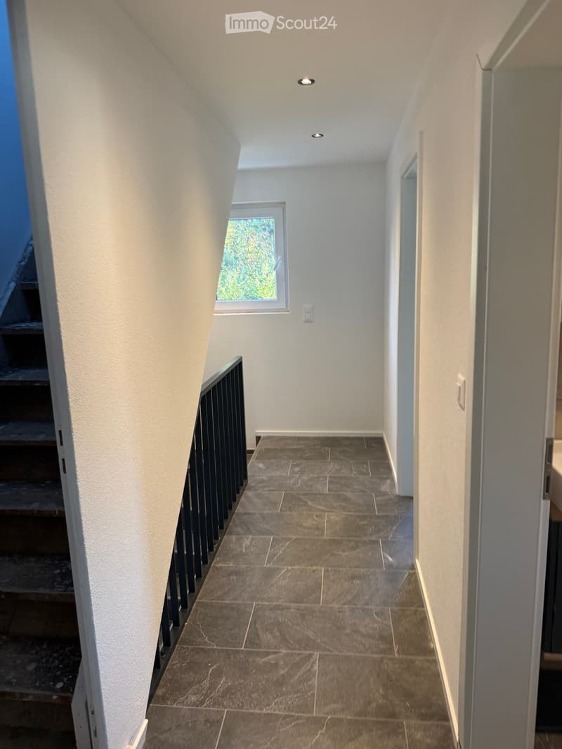 Modern hallway with tiled floor, white walls, and recessed lighting. Features a window with a view of greenery and a staircase with a black railing visible to the left.