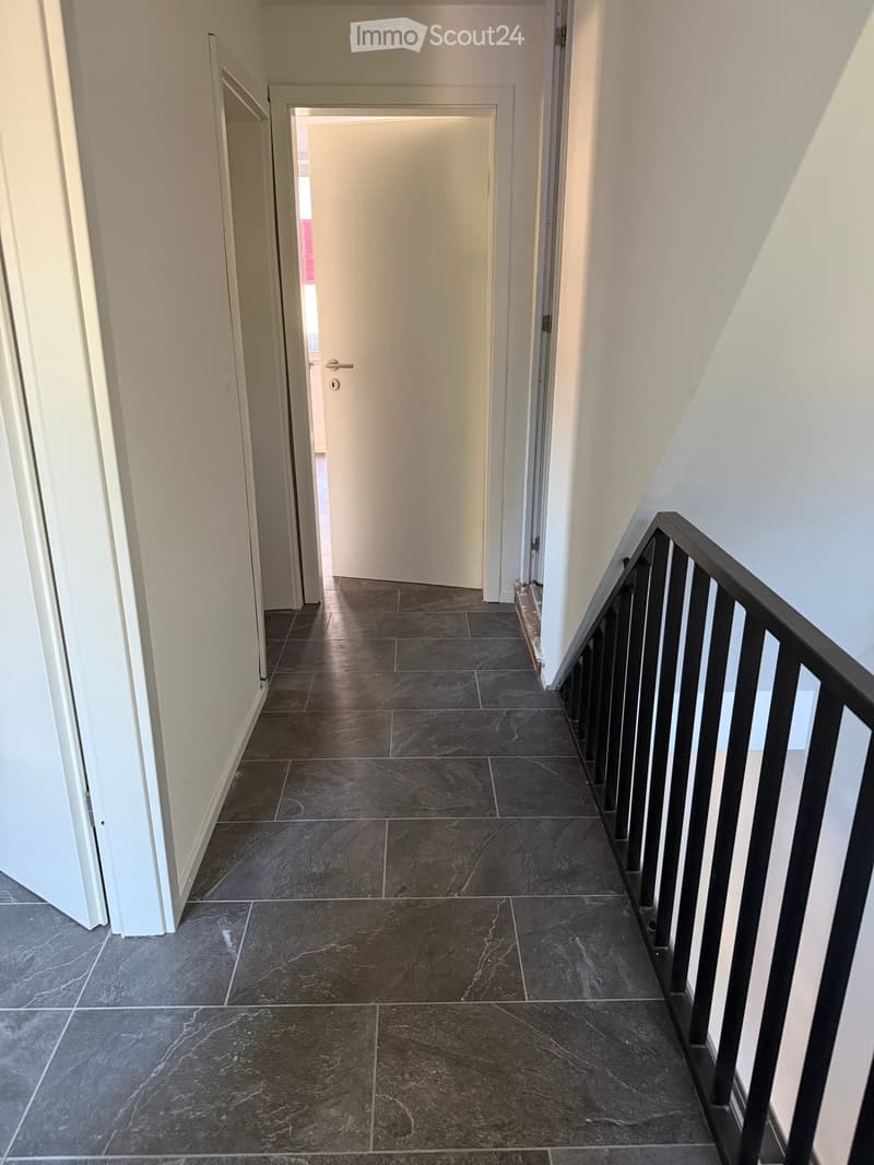A narrow hallway with dark tiled flooring, white walls, and a black metal staircase railing on the right side. A closed white door is visible at the end of the hallway.