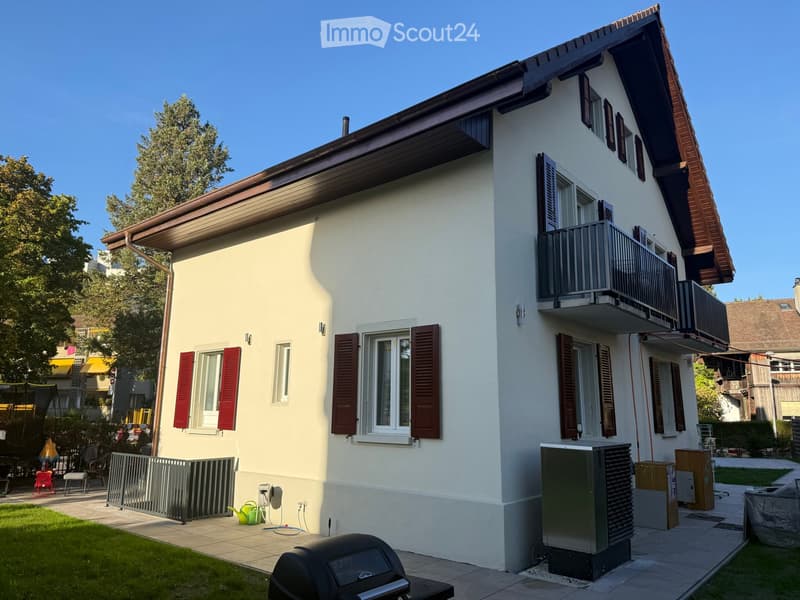 Two-story house with white walls, red shutters, and a balcony on the upper floor. The house has a well-maintained garden with a barbecue, a small fence, and outdoor furniture.