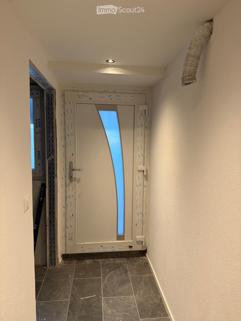 Empty hallway with tiled floor, white walls, and a partially open door leading to another room. The door has a glass panel and a visible ventilation duct on the ceiling.