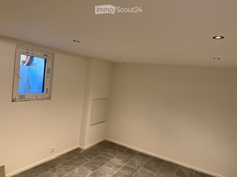 Empty room with beige walls, dark tiled floor, and a single window with a white frame. The ceiling has recessed lighting.