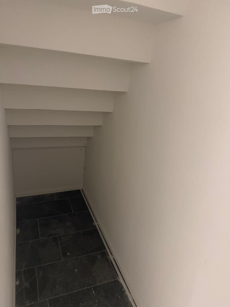 A narrow hallway with white walls and black tiled flooring, featuring a built-in shelf on one wall.