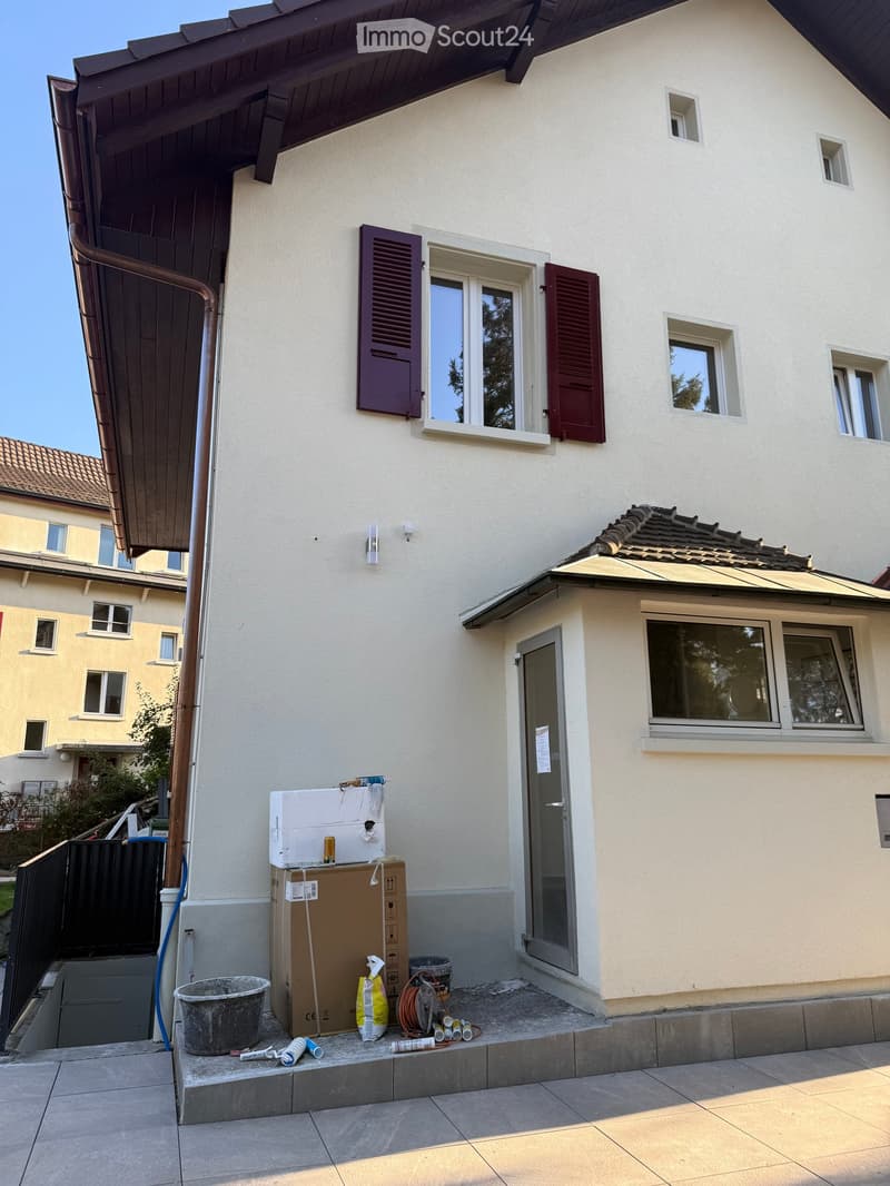 Two-story house with white exterior, multiple windows, and a small entrance area with construction materials and tools.