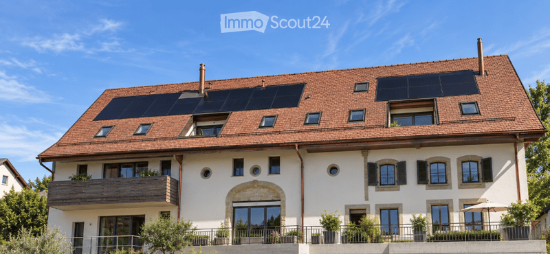 A two-story house with a red-tiled roof, white exterior walls, multiple windows, and solar panels on the roof. The house features a balcony on the first floor and is surrounded by greenery.