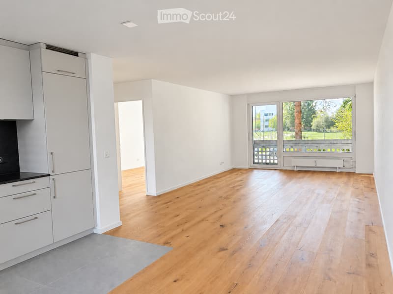 Empty room with light wooden flooring, white walls, and a modern kitchen area featuring white cabinets and a black countertop. Large windows offer a view of greenery outside.