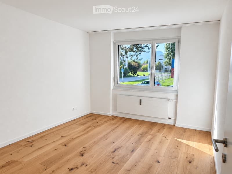 Empty room with light wooden flooring, white walls, and a large window with a view of the outside. A radiator is mounted below the window.