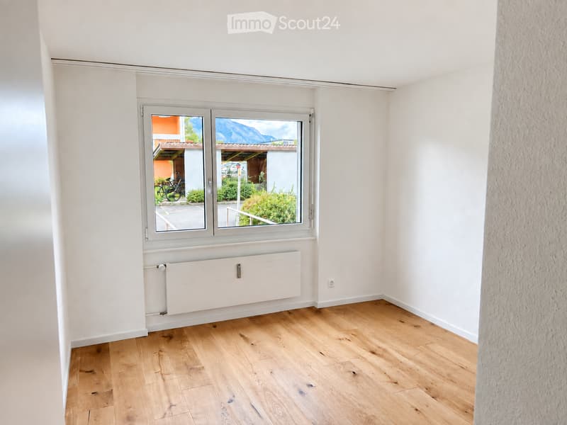 Empty room with light wooden flooring, white walls, and a large window offering a view of the outdoors.