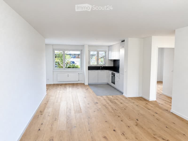 Bright, minimalist kitchen with white cabinets, a sink, and an oven, featuring light wooden flooring and large windows offering a view of greenery.