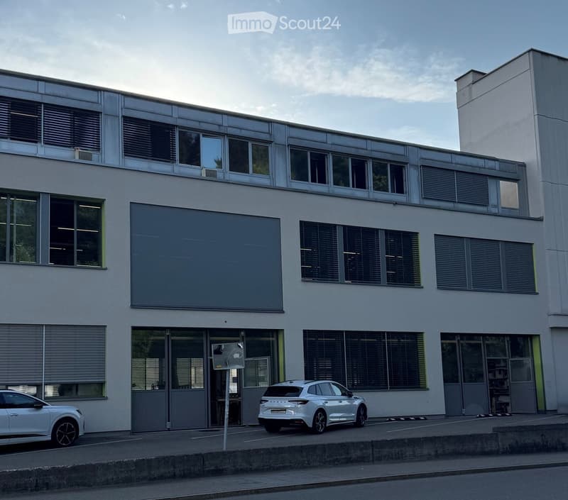 Modern two-story building with large windows, grey facade, and a large blank wall on the first floor. Two cars are parked in front.