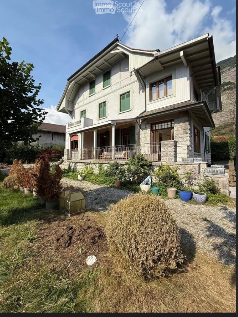 Two-story house with white siding, green shutters, and a stone base. Features a covered porch with railings and several windows. The front yard includes potted plants and a small garden area.