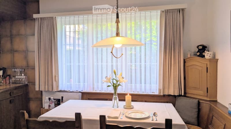 Dining room with a table set for two, a hanging lamp, a vase with flowers, and a small cabinet with a decorative item on top.