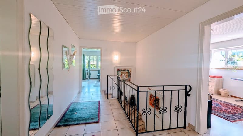 Bright hallway with white walls, tiled floor, and a staircase with a black wrought-iron railing. Mirrors, artwork, and a small rug are visible.