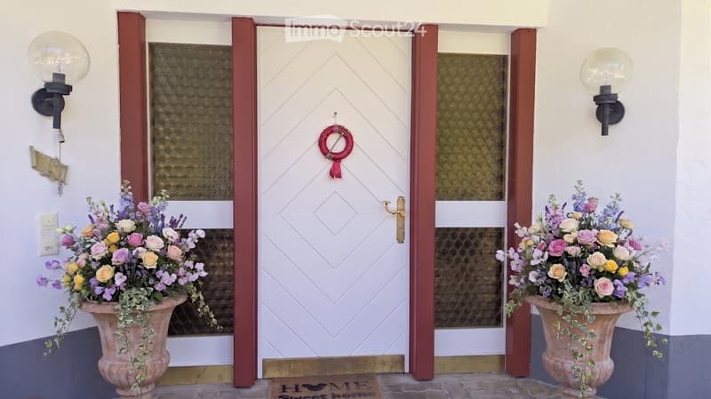 Entrance area with a white double door flanked by two large flower arrangements in urns, decorative wall sconces, and a "Sweet Home" doormat.