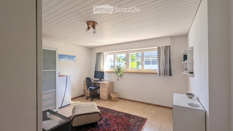 Bright, modern room with wooden flooring, large windows, a desk with a computer, a comfortable chair, a small table, and a shelf with books.
