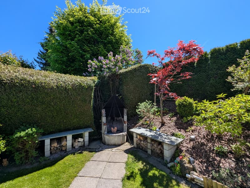 Well-maintained garden with a stone fireplace, neatly stacked firewood, benches, and various trees and shrubs.