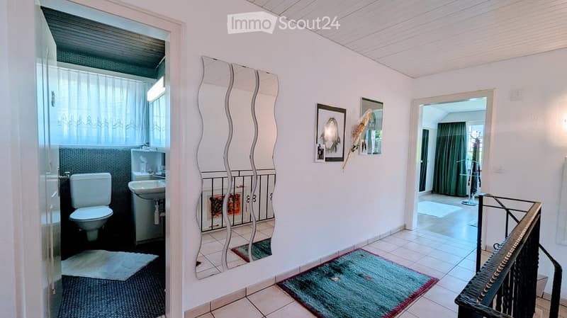 A well-lit hallway with white walls, tiled flooring, and a decorative mirror. It features a small rug, a staircase leading upwards, and an open doorway to a bathroom area with a toilet and sink.