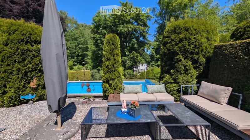 Well-maintained garden with a swimming pool, patio furniture including a sofa, coffee table, and chairs, surrounded by lush greenery and trees.